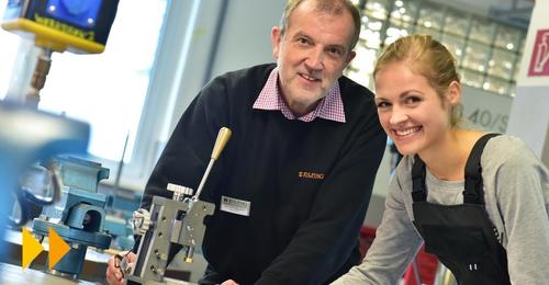 Ein Mann und eine Frau, beide lächelnd, arbeiten gemeinsam in einer Werkstatt, wobei der Mann ein Werkzeug hält und die Frau assistiert, umgeben von Werkzeugen und Geräten.